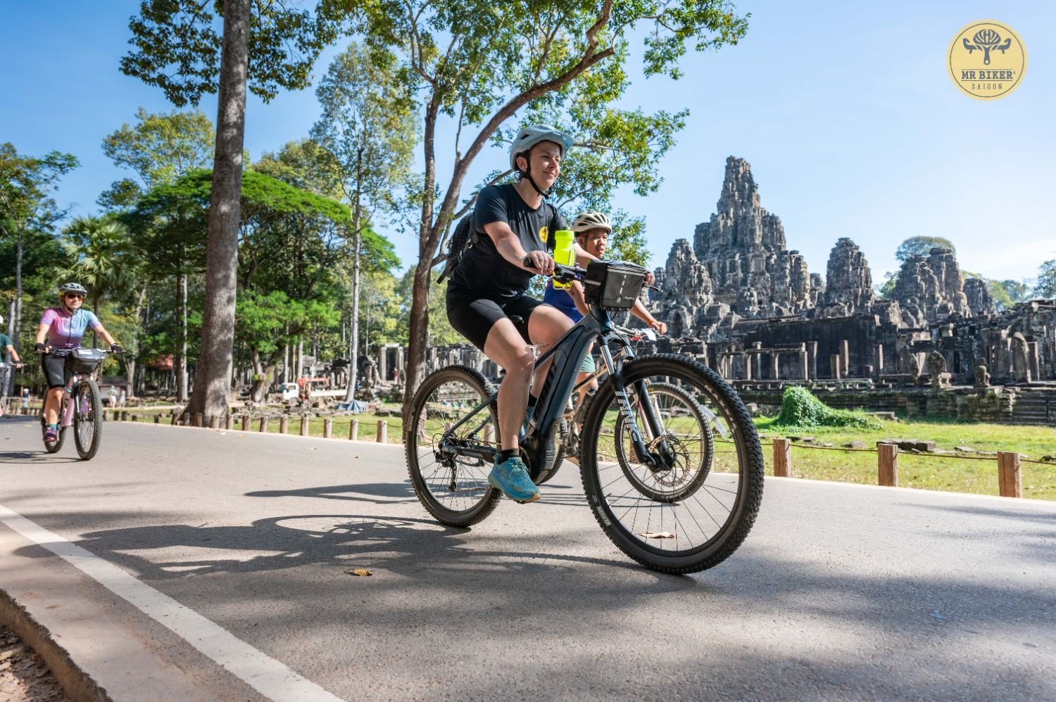Cyclists riding past the carved face towers of Bayon Temple in Angkor Thom, Siem Reap, Cambodia